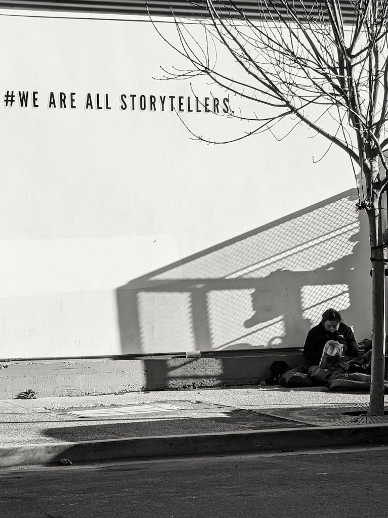 Monochrome photo of a person sitting on a sidewalk under 'We Are All Storytellers' graffiti.