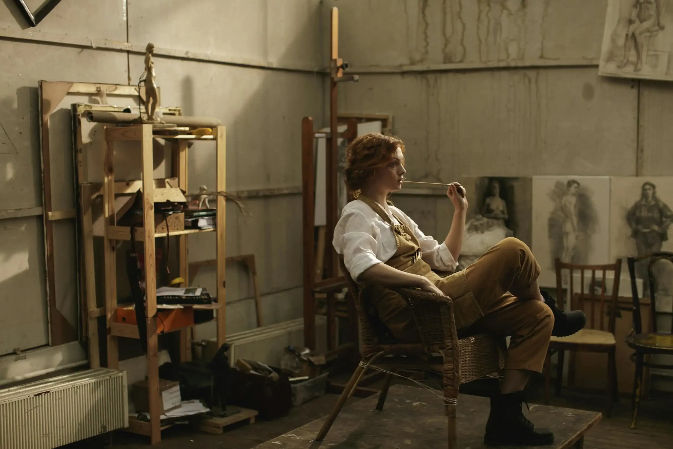 Female artist in a lofty art studio, lost in thought with a paintbrush, surrounded by drawings.